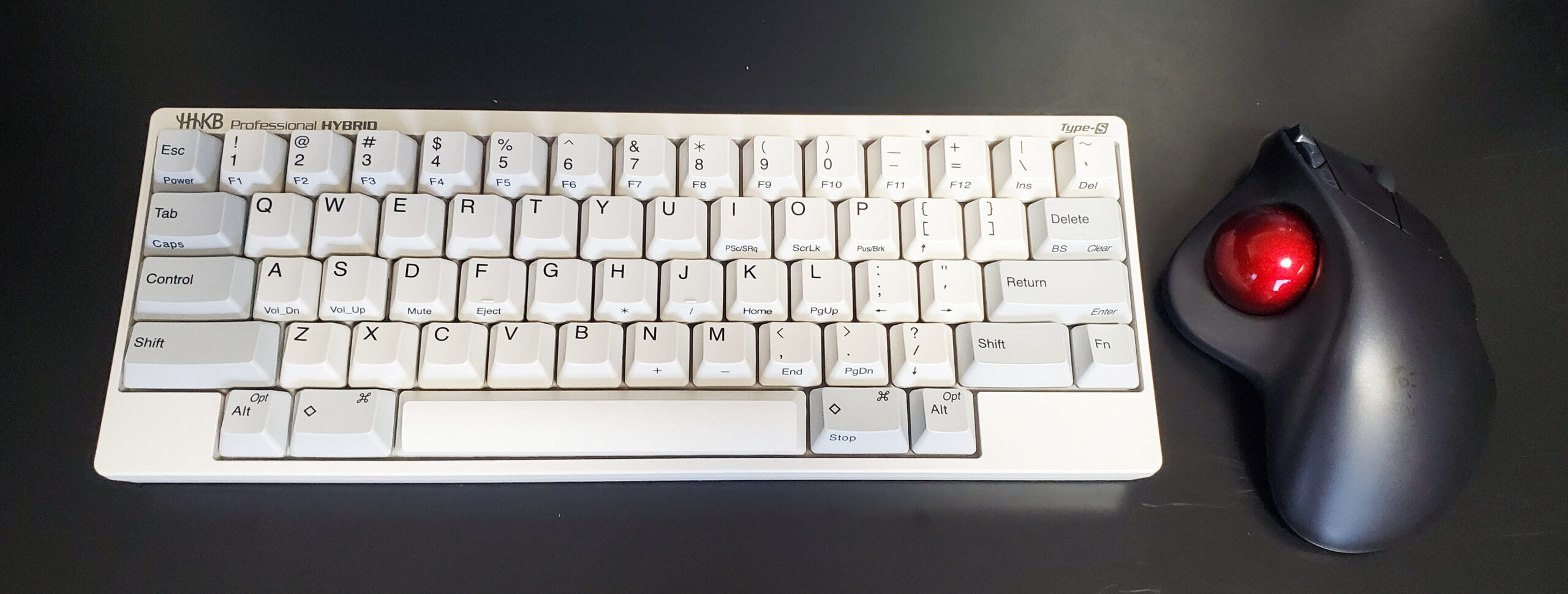 黒いデスクに白キーキャップのHHKBと赤いボールのトラックボールを並べた作業環境